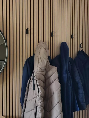 Een houten lattenwand is voorzien van vier yessential.nl Huso Black haken, die elk een beige of blauwe jas of jack vasthouden. Aan de linkerkant van de muur hangt een ronde spiegel.