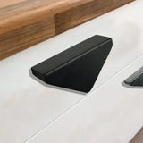 De Triano Black 64mm handgreep van yessential.nl, met een modern driehoekig ontwerp, is gemonteerd op een wit ladefront onder een houten aanrechtblad.