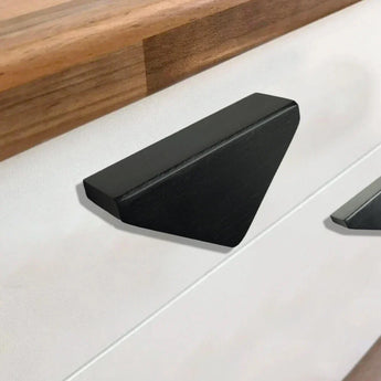 De Triano Black 64mm handgreep van yessential.nl, met een modern driehoekig ontwerp, is gemonteerd op een wit ladefront onder een houten aanrechtblad.