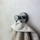 Een close-up laat zien hoe de Mano Gunmetal WC set van yessential.nl een beige handdoek met textuur vasthoudt, gemonteerd op een lichte muur - een elegante upgrade van je moderne toiletaccessoires.