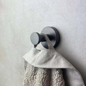 Een close-up van de Kyano Gunmetal Hook van yessential.nl die een beige handdoek met textuur aan zijn lus vasthoudt, gemonteerd op een lichtgekleurde muur.
