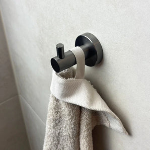 De Justo Gunmetal Haak van yessential.nl houdt een witte handdoek op een lichtgrijze betegelde muur en voegt minimalistische elegantie toe aan elke moderne badkamer.