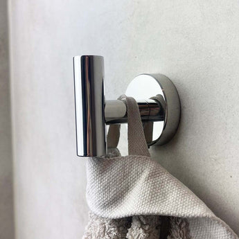 Een close-up van de Sandro Chroom WC-set van yessential.nl, met een glanzend chromen haakje dat een beige handdoek met textuur vasthoudt tegen een lichte muur, wat elegantie brengt in je badkameraccessoires.
