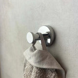 Een beige handdoek met lus hangt aan een zilveren wandhaakje, onderdeel van de Xavo Chroom Wc-set van yessential.nl, tegen een gladde, lichtgekleurde muur.