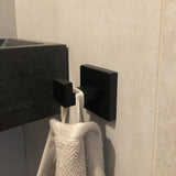 Een close-up van de Valo Black Hook van yessential.nl die een handdoek met beige textuur vasthoudt, gemonteerd op een lichte badkamermuur naast een donkere wastafel.