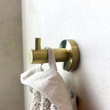 Een close-up van de Justo Gold Hook van yessential.nl die een beige handdoek met textuur vasthoudt tegen een lichte muur. De haak heeft een moderne, cilindervormige vorm en een matgouden afwerking.