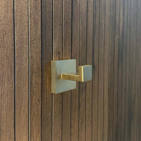 De Valo Gold Hook van yessential.nl, een goudkleurige handdoekhaak met vierkante basis, is gemonteerd op een donkerbruine houten stok met smalle panelen en is ideaal als handdoekhaken badkamer.