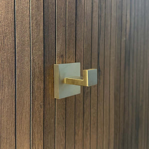 De Valo Gold Hook van yessential.nl, een goudkleurige handdoekhaak met vierkante basis, is gemonteerd op een donkerbruine houten stok met smalle panelen en is ideaal als handdoekhaken badkamer.