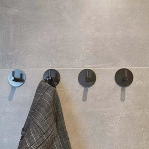 Vier Rudro Gunmetal wandhaken van yessential.nl zijn op een rij gemonteerd op een grijs betegelde muur. Aan de tweede haak hangt een donker kledingstuk met patroon, terwijl de andere drie leeg blijven.