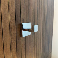 Een Chromen haak met een vierkante rozet, is gemonteerd op een verticaal houten paneel met een donkerbruine gegroefde textuur.