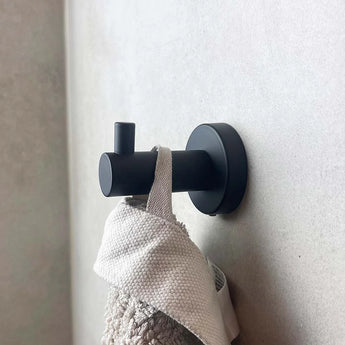 De Justo Black Hook van yessential.nl, met een modern cilindervormig ontwerp, houdt een beige handdoek met textuur tegen een lichte muur en voegt zowel stijl als functie toe aan je badkamer.
