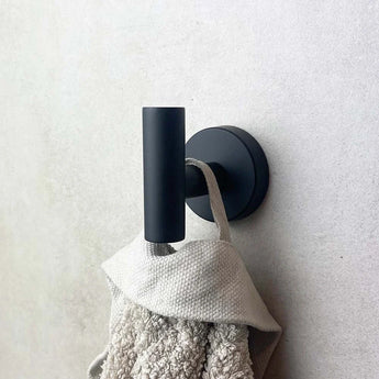 De Sandro Black WC-set van yessential.nl heeft een zwart metalen wandhaakje dat een handdoek met beige textuur tegen een lichtgekleurde muur houdt.