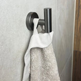 Een Rodrigo Gunmetal haak van yessential.nl is gemonteerd op een betegelde badkamermuur en toont stijlvol een keurig gedrapeerde witte handdoek.