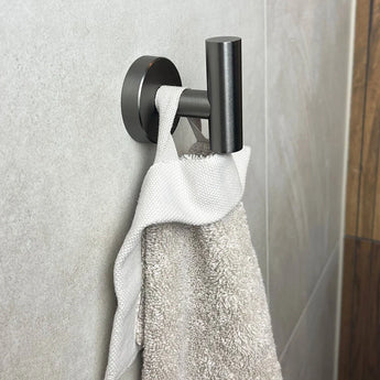 Een Rodrigo Gunmetal haak van yessential.nl is gemonteerd op een betegelde badkamermuur en toont stijlvol een keurig gedrapeerde witte handdoek.