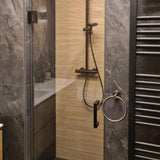 Een moderne douche met Elarbo Gunmetal armaturen van yessential.nl, een glazen deur, verticale beige structuurmuur, donkergrijze stenen tegels, ronde handdoekring badkameraccessoires en een zwarte handdoekradiator rechts.