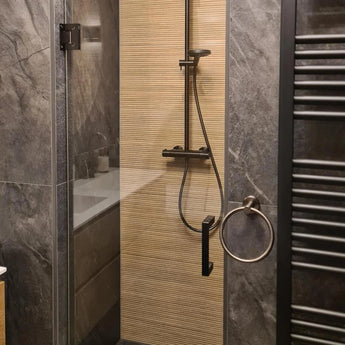 Een moderne douche met Elarbo Gunmetal armaturen van yessential.nl, een glazen deur, verticale beige structuurmuur, donkergrijze stenen tegels, ronde handdoekring badkameraccessoires en een zwarte handdoekradiator rechts.