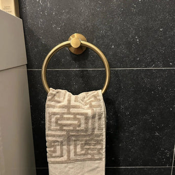 Een handdoek met beige patroon hangt aan de Elarbo Gold van yessential.nl, een stijlvolle gouden handdoekring gemonteerd op een donkere, stenen badkamermuur.
