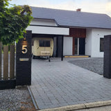 Een modern wit huis met een donker dak is voorzien van het yessential.nl Angelo RVS 20cm huisnummer 5 op strakke zwarte hekpijlers. Een kleine beige caravan staat onder de carport en een lommerrijke boom bij de ingang zorgt voor extra charme.