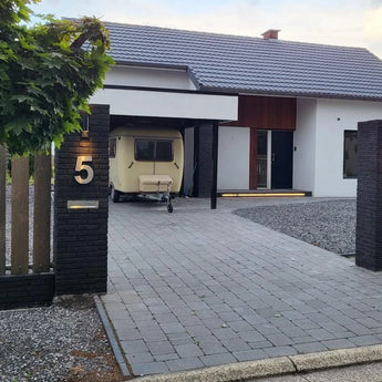 Een modern wit huis met een donker dak is voorzien van het yessential.nl Angelo RVS 20cm huisnummer 5 op strakke zwarte hekpijlers. Een kleine beige caravan staat onder de carport en een lommerrijke boom bij de ingang zorgt voor extra charme.