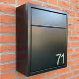 Een moderne zwarte brievenbus met het nummer 71, gemaakt met Animo RVS 7,6cm (geen boor) plaknummers van yessential.nl, is gemonteerd op een rode bakstenen muur.