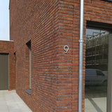 Een modern bakstenen gebouw met een groot raam en deur toont een yessential.nl Milano RVS 15cm huisnummer in geborsteld roestvrij staal op de hoekmuur. Naast het raam loopt een metalen regenpijp die een bestelbus weerspiegelt.