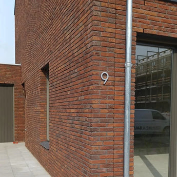 Een modern bakstenen gebouw met een groot raam en deur toont een yessential.nl Milano RVS 15cm huisnummer in geborsteld roestvrij staal op de hoekmuur. Naast het raam loopt een metalen regenpijp die een bestelbus weerspiegelt.