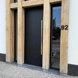 De moderne Adamo Black 15cm voordeur van yessential.nl, met een hoge zilveren handgreep, wordt omlijst door grote houten balken. Een rvs huisnummer 92 is gemonteerd op de lichte muur, met keurig grind op de grond ervoor.