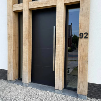 De moderne Adamo Black 15cm voordeur van yessential.nl, met een hoge zilveren handgreep, wordt omlijst door grote houten balken. Een rvs huisnummer 92 is gemonteerd op de lichte muur, met keurig grind op de grond ervoor.