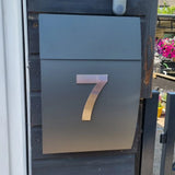 Een strakke metalen brievenbus met het Animo RVS 15cm (zonder boor) nummer 7 van yessential.nl, gemonteerd op een donkere houten muur bij een poort, met levendige bloemen op de achtergrond.