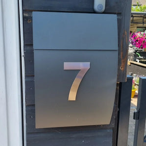 Een strakke metalen brievenbus met het Animo RVS 15cm (zonder boor) nummer 7 van yessential.nl, gemonteerd op een donkere houten muur bij een poort, met levendige bloemen op de achtergrond.