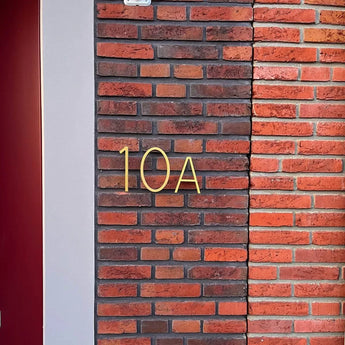 De Amato Gold 15cm nummer en letter van yessential.nl zijn vormgegeven als moderne huisnummers en gemonteerd op een bruin en rode bakstenen muur naast een lichtgrijze verticale strip en een rood deurkozijndeel aan de linkerkant.