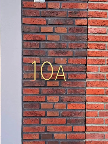 De Amato Gold 15cm nummer en letter van yessential.nl zijn vormgegeven als moderne huisnummers en gemonteerd op een bruin en rode bakstenen muur naast een lichtgrijze verticale strip en een rood deurkozijndeel aan de linkerkant.