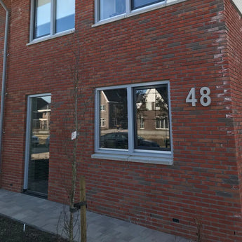 Een bakstenen gebouw met een yessential.nl Enrico beton 30cm huisnummer op de muur, met twee grote ramen en een kleine boom op de geplaveide stoep. Reflecties van naburige huizen verschijnen in de ramen.