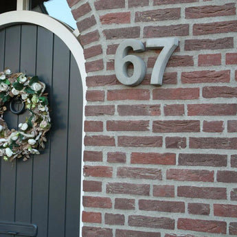 Een close-up van een bakstenen muur toont het Enrico beton 20cm huisnummer 67 van yessential.nl in grote metalen cijfers, gemonteerd naast een donkergrijze boogdeur versierd met een groen-witte bloemenkrans.