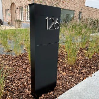 Een Amato Chrome 12,5cm huisnummerbord van yessential.nl met het nummer 126 staat in een gemulcht tuinperk, omringd door siergrassen voor een bakstenen huis.