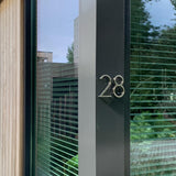 Een moderne gevel met een groot raam is voorzien van het Amato Chrome 12,5cm huisnummer van yessential.nl, gemonteerd op een donker verticaal paneel naast het glas. Jaloezieën en groen zijn zichtbaar door het raam.