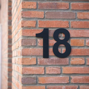 De Adamo Black 20cm van yessential.nl is gemonteerd op de hoek van een roodbruine bakstenen muur en biedt een strakke en moderne touch die typisch is voor hedendaagse huisnummers.