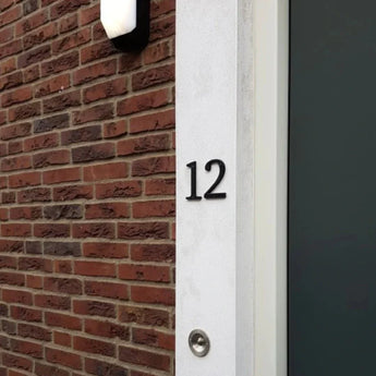 Huisnummer 12, gemaakt met de Matteo Black 10cm van yessential.nl, is gemonteerd op een witte muur naast bakstenen. Een deurbel bevindt zich onder het nummer en een donkere deur verschijnt gedeeltelijk aan de rechterkant.