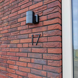 Een moderne zwarte wandlamp en yessential.nl Amato Black huisnummers van 15 cm, waaronder nummer 17, staan op een rode bakstenen muur naast een wit omlijst raam.