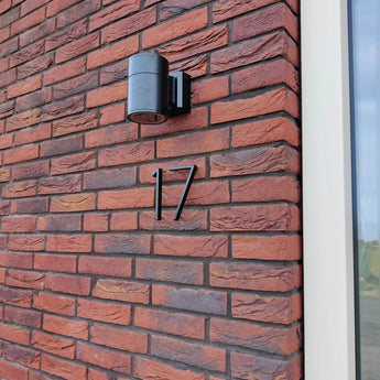 Een moderne zwarte wandlamp en yessential.nl Amato Black huisnummers van 15 cm, waaronder nummer 17, staan op een rode bakstenen muur naast een wit omlijst raam.