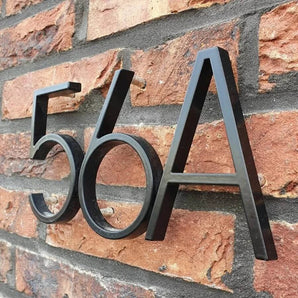 De Amato Black 12,5cm huisnummers van yessential.nl zijn gemonteerd op een rode bakstenen muur in een moderne schreefloze stijl, waarbij de verhoogde cijfers en letters opvallende schaduwen achter zich werpen.