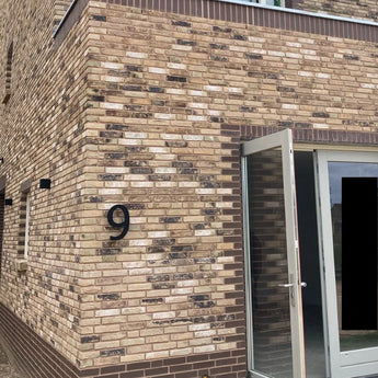 In een bakstenen gebouw is een yessential.nl Carlo Black 20cm 3D huisnummer 9 gemonteerd bij een gedeeltelijk openstaande glazen deur, waarbij de muur bruin en taankleurig metselwerk vertoont.