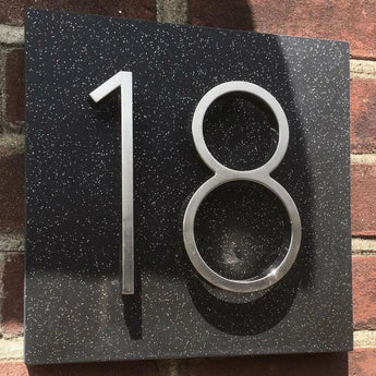 De Amato Chrome 12,5cm van yessential.nl is een metalen huisnummer in een verchroomde stijl, gemonteerd op een zwart gespikkelde plaquette op een bakstenen muur. Zonlicht werpt schaduwen die de driedimensionale look accentueren.