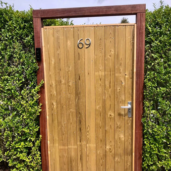 Een tuinpoort met het yessential.nl Matteo Nickel 10cm huisnummer "69" op donker hout, omlijst door hoge hagen. Een zilveren handgreep en toetsenbord voegen een geborsteld accent toe aan de rechterkant van de poort.