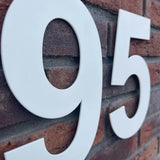 Grote witte huisnummers "95" in de stijl Adamo White 20cm van yessential.nl zijn gemonteerd op een rode bakstenen muur. De schuine foto benadrukt zowel de baksteen textuur als de gladde afwerking van deze huisnummer wit.