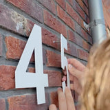 Iemand installeert een yessential.nl Adamo White 20cm huisnummer naast een witte 4 op een rode bakstenen muur. Een deel van een metalen pijp verschijnt aan de rechterkant.