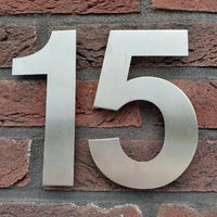De Adamo RVS 15cm van yessential.nl is een groot metalen huisnummer 15, elegant ontworpen voor montage op een rode bakstenen muur - perfect voor wie op zoek is naar stijlvolle, duurzame huisnummers.