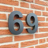 De Angelo Black 20cm van yessential.nl zijn grote zwarte 3D huisnummers, gemonteerd op een rood bakstenen muur met zichtbare voegen en rustieke tekstuur, ideaal als moderne huisnummers.