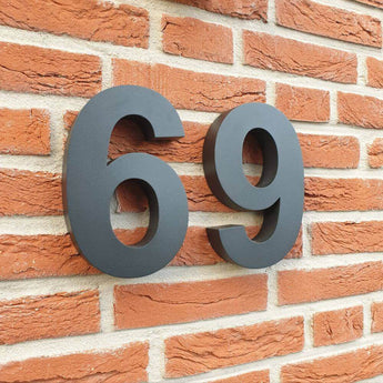 De Angelo Black 20cm van yessential.nl zijn grote zwarte 3D huisnummers, gemonteerd op een rood bakstenen muur met zichtbare voegen en rustieke tekstuur, ideaal als moderne huisnummers.