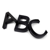 Drie grote, zwarte, 3D letters-A, B en C-Carlo Black 20cm by yessential.nl-liggen op een wit oppervlak, overlappen elkaar een beetje en staan onder verschillende hoeken als moderne huisnummers.